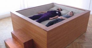 Two people laying in a wooden box shaped bed 