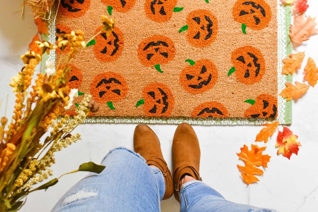 jack o lantern door mat on the ground with fall colored leaves