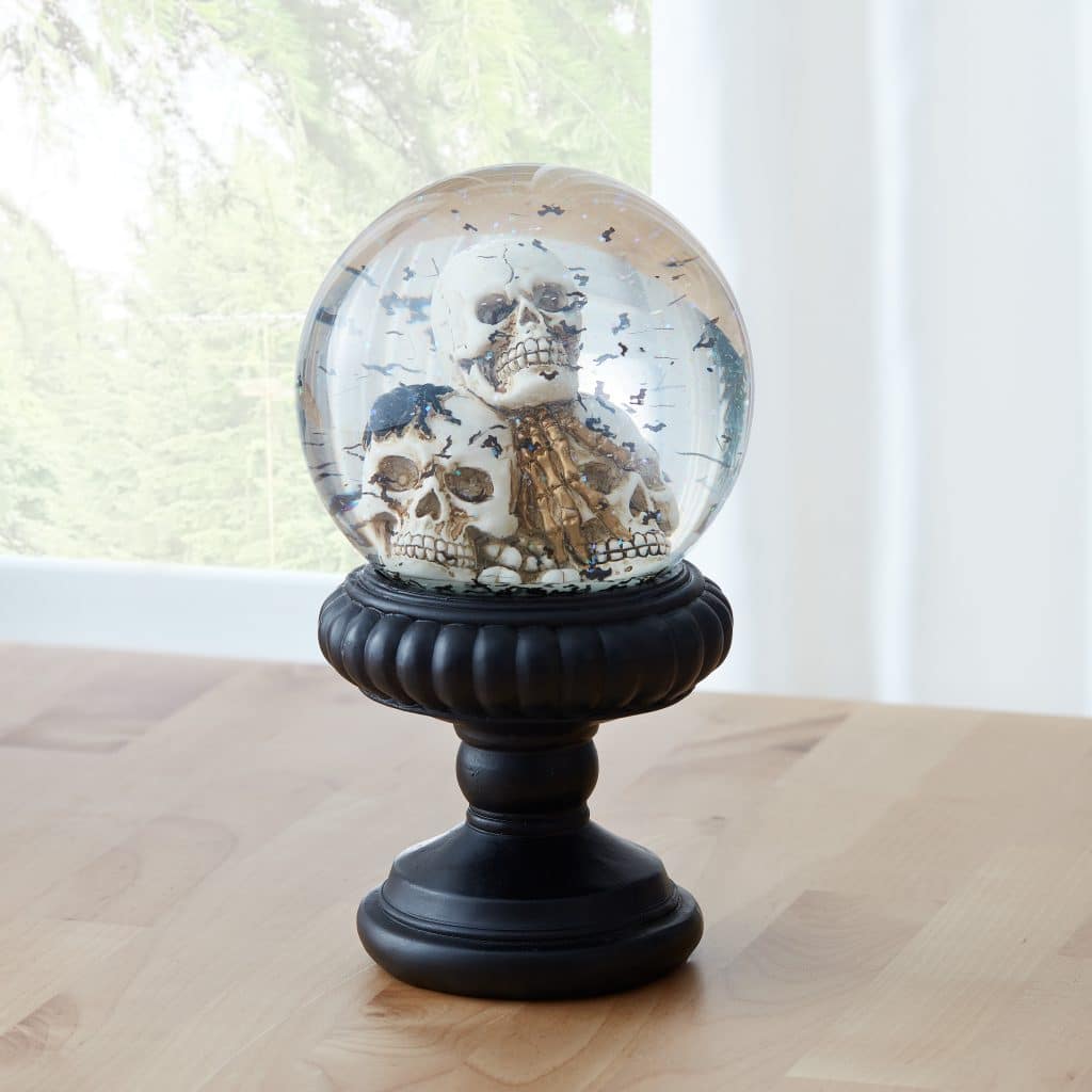 two skulls and small black bats inside a snowglobe with a black holder undeneath on a wooden table next to a window